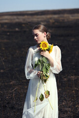 Young woman in white dress honding sunflowers stands on black burnt field