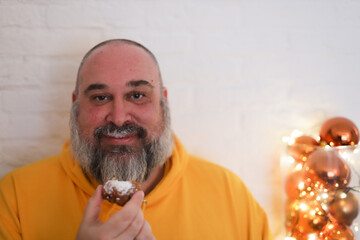 Man with grey beard in yellow hoodie eats and enjoys fried donut with sugar powder.