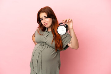 Fototapeta premium Young redhead caucasian woman isolated on pink background pregnant and holding clock while thinking