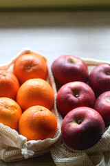 Two sustainable shopping bags filled with apples and oranges on wooden table. Selective focus.
