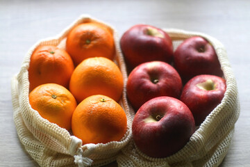 Two sustainable shopping bags filled with apples and oranges on wooden table. Selective focus.