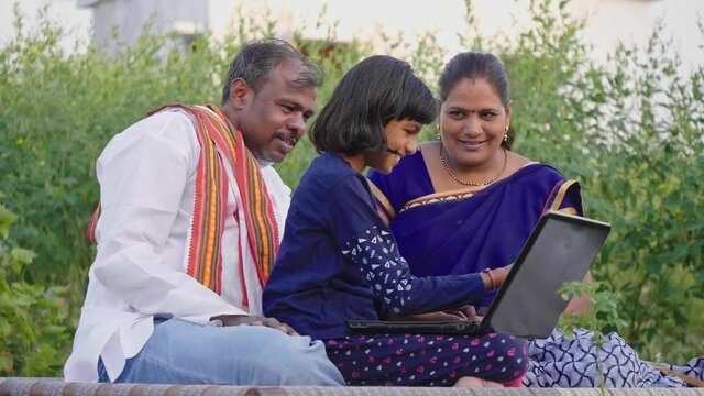 Happy Indian farmer family sitting outdoors on the farm including father, mother, and daughter using a laptop and interacting with each other. Concept of rural Family, relationship and farming