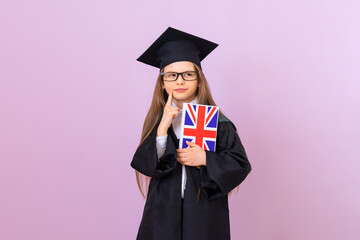 A young graduate in a ceremonial raincoat and a square graduation hat holds an English language book and looks thoughtfully ahead. a schoolgirl on an isolated background.