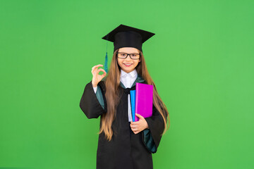 A beautiful and joyful graduate in a graduation cap and a ceremonial robe, holds books and shows a hand gesture everything is OK. isolated green background.