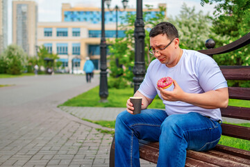 a despondency, puzzled, sad man with glasses is sitting in a city park, having lunch, eating a sweet donut, a bun and holding a cup of coffee, tea