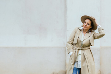 Fototapeta na wymiar Woman in stylish trench coat and hat looking at camera while posing outdoors on the street.