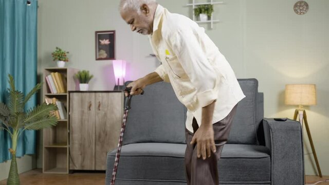 Old man using walking stick as support for walking while sitting on sofa at home - concept of seniors knee joint paint, suffering from Osteoarthritis and arthritis