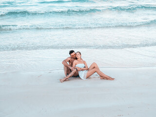 Handsome man and pretty woman sitting on the beach with the sea background and looking into the distance. Outdoors, closeup. Concept of leisure and travel