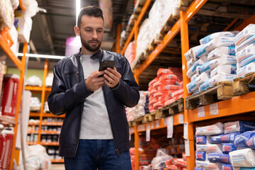 a man at a hardware store looking at a phone screen next to shelving with bags of putty and plaster.