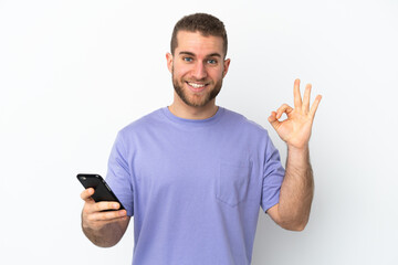 Young handsome caucasian man isolated on white background using mobile phone and doing OK sign