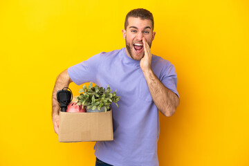 Young caucasian making a move while picking up a box full of things isolated on yellow background shouting with mouth wide open