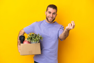 Young caucasian making a move while picking up a box full of things isolated on yellow background making money gesture