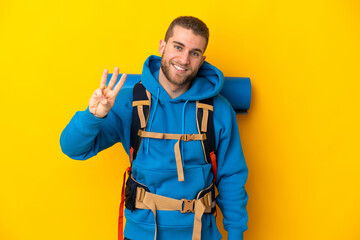 Young caucasian mountaineer man with a big backpack isolated on yellow background happy and counting three with fingers