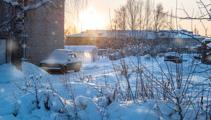 Winter evening at snowy street in village. The rays of the setting sun illuminate buildings,...