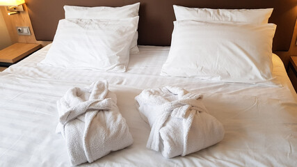 2 snow-white bath robes, beautifully folded, lie on a bed covered with white linen.