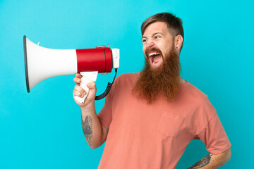 Young reddish caucasian man isolated on blue background holding a megaphone and smiling