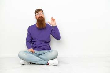 Young caucasian reddish man sitting on the floor isolated on white background with fingers crossing and wishing the best