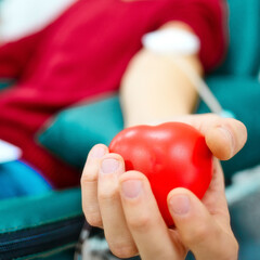 Topic of donation. Man donates blood in hospital. Man's hand squeezes rubber heart. Close-up. Donor sits in chair. Background. - obrazy, fototapety, plakaty