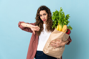 Young woman holding a grocery shopping bag isolated on blue background showing thumb down with...
