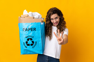 Young woman holding a recycling bag full of paper to recycle isolated on yellow background happy and counting three with fingers