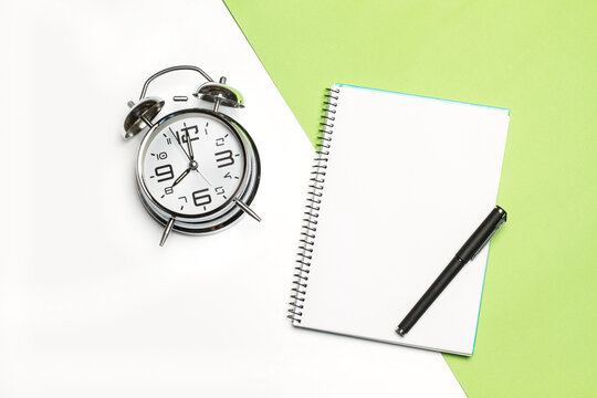 Cuaderno en blanco para escribir con espirales junto a un reloj despertador y un boligrafo sobre un fondo blanco y verde liso y aislado. Vista superior. Copy space