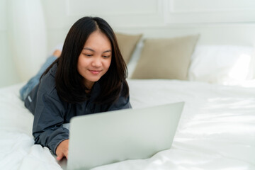 Portrait, a cute Asian teenage girl. wearing gray shirt lying in bed with a notebook computer in the bedroom to study online with a happy face