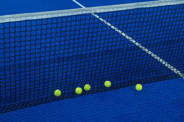 Some balls next to the net of a blue paddle tennis court.