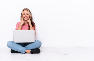 Young Russian girl with a laptop sitting on the floor isolated on white background smiling with a happy and pleasant expression