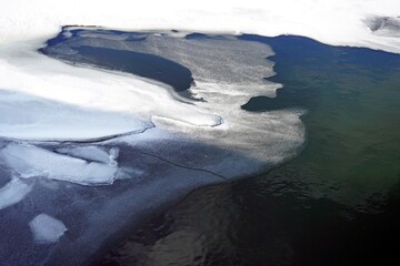Winter. January. Ice and thawed patches on the surface of the lake. White snow. Deep water color.