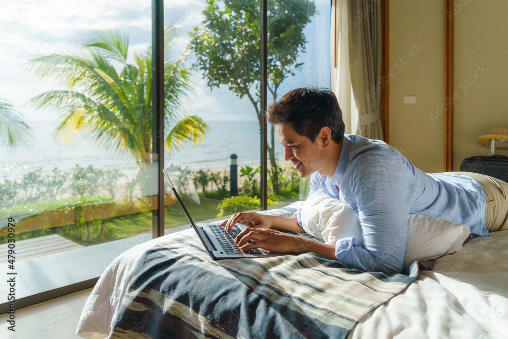 Wall mural Young Asian businessman lying in bed and working with laptop in room at resort near sea during a summer vacation holiday travel..