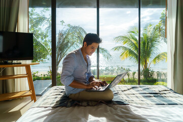 Young Asian businessman sitting in bed and working with laptop in room at resort near sea during a...