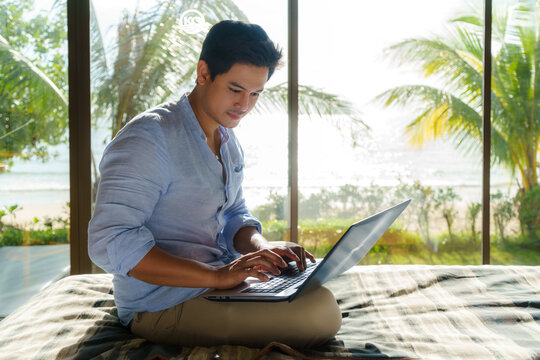 Young Asian businessman sitting in bed and working with laptop in room at resort near sea during a summer vacation holiday travel..