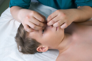 Fototapeta na wymiar Session of craniosacral therapy, cure of teen boy's nose by doctor therapist. Craniosacral therapist touches the boy's nose and checks corrects of the nose by hands at hospital, side view of face.