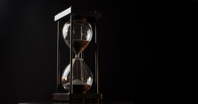 Rotation of sand timer clock with sand flowing from through hourglass on black background.
