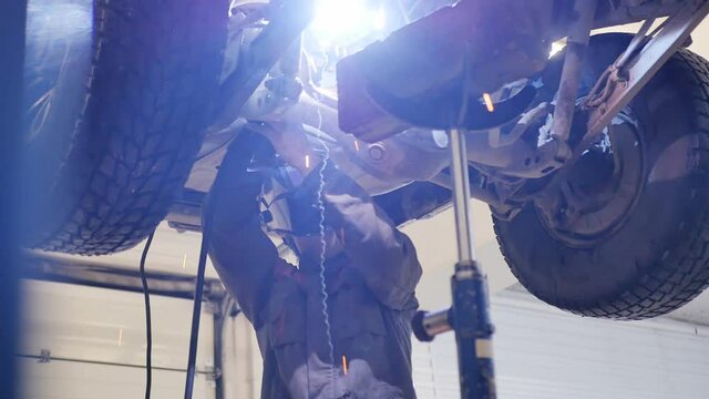 a man gas-electric welder welds a part of the rear suspension of an SUV by lifting the car up with a lift and standing under it. He takes off the mask and looks at the result