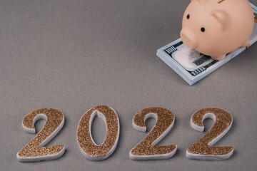 Piggy bank on a bundle of hundred-dollar bills and Number 2022 on a gray background. Copy space