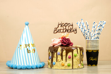 Birthday layer cake decorated with chocolate pieces and happy birthday text, hazelnuts and roses flowers and disposable cups and straws and blue hat party on wood table