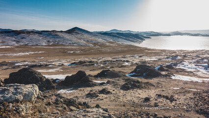 The frozen lake is surrounded by snow-covered hills. Boulders are scattered on the bare ground. A sunny winter day in Siberia. Blue sky. Baikal