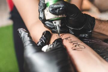 Fototapeta A professional tattoo artist uses a rotary tattoo machine with a round liner cartidge to draw cursive names on a female client's forearm. obraz
