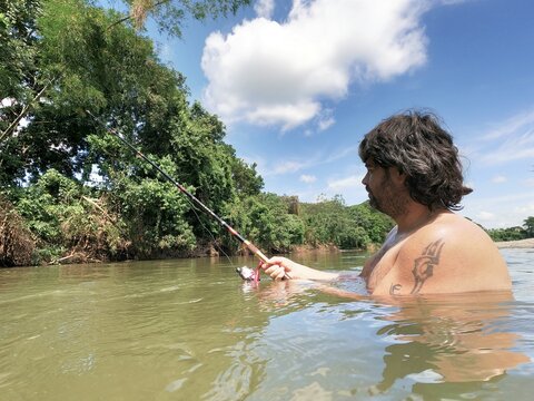 Hombre pescando en rio con mucha vegetación de fondo, naturaleza verde, cielo azul y nubes blancas