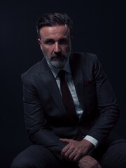 Portrait of adult businessman wearing trendy suit and sitting in modern studio on stylish chair against the black background