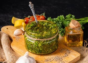 Glass jar with green chimichurri with spoon inside home made on wooden board, dark background.