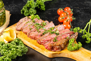 Beef steak cooked and cut into pieces, served on a wooden board and sprinkled with green chemichurri sauce, dark table on a background of tomatoes, parsley.