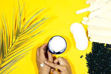 depilatory pearly blue solid wax beans and wooden stick, cotton pads and woman making hand cream box and palm leaf on yellow background