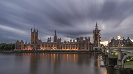 Naklejka premium The Houses of Parliament, London, England, illuminated as night falls and reflecting on the river Thames, with clouds streaking overhead
