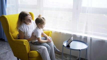 Parent and child in pagamas spending morning together. Cheerful blonde mommy with loose hair and funny toddler daughter watch cartoons on tablet in bright yellow armchair near big window. 