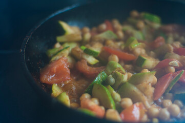 Close-up of juicy vegetables with legumes stewed in a pan surrounded by a dark background. Zucchini, tomatoes, chickpeas. Cooking a healthy vegetarian meal soft focus.