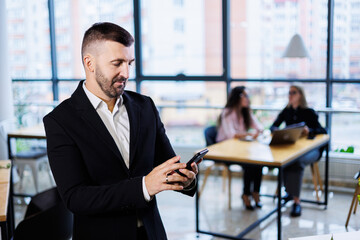 Portrait of stylish young businessman standing in large modern office high on top floor and looking at phone