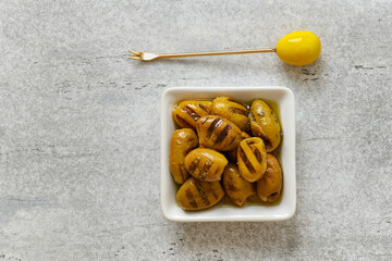 Grilled olives barbecued in olive oil with garlic in a square bowl and olive fork, top view