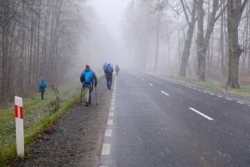 Fototapeta premium Little snowy asphalt road. Silhouettes of people walking by side of road in snowy misty day.